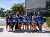A group of women wearing blue shirts with "USA" in white lettering smile as they pose together outdoors, with a construction facade, trees and shrubs in the background.