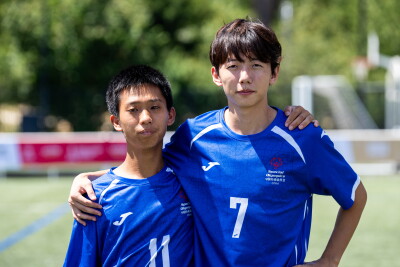Dos compañeros de equipo de fútbol, vestidos con camisetas azules de las Olimpiadas Especiales de China, están de pie uno al lado del otro en un campo de fútbol, abrazados por los hombros. El jugador de la izquierda lleva el número 11, mientras que el de la derecha lleva el número 7. Al fondo, borroso, se aprecian árboles y una portería.