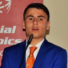 Gilmour Borg in a blue suite and red tie standing speaking behind a podium and in front of the Special Olympics logo backdrop.