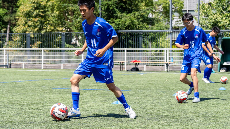 A football player in a blue number 11 jersey dribbles a ball through a training drill on an artificial turf field while teammates complete similar exercises in the background.