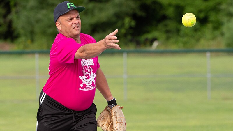 Kevin stands staring after the softball he just threw with his right hand. His hand is still extended. He has a softball mitt on his left hand.