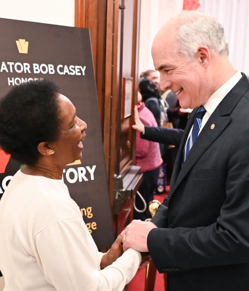 Loretta and Senator Casey shake hands standing next to the Black History event sign. 