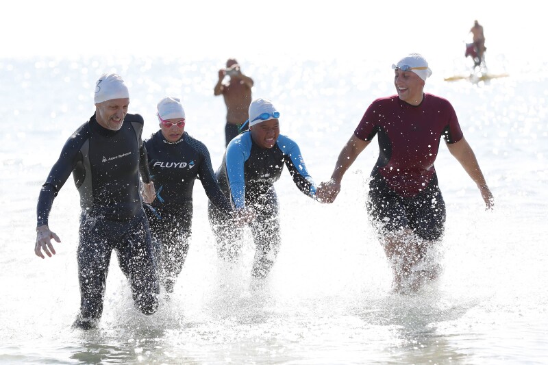 A man and three women wearing wet suits and swim hats and goggles on their heads, hold hands and smile as they walk out of the sea together with water splashing around their legs.
