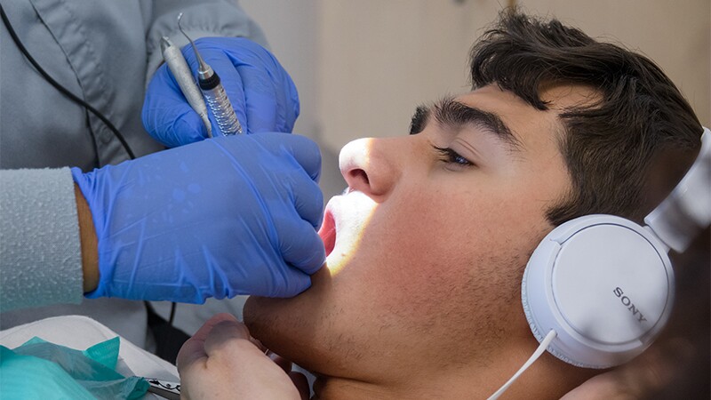 A male athlete has his teeth cleaned by a dental technician. He is listening to music on his headphones.