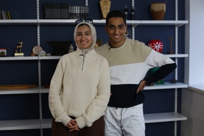 Young man and woman standing in front of a bookshelf with nicknacks on it. 