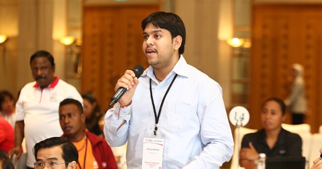 Man speaking on a microphone in a room of round tables with people at them.