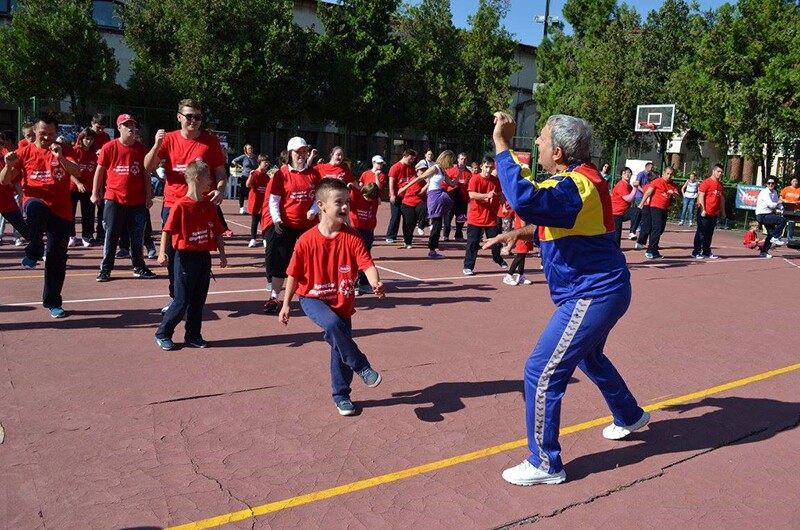 Coach and athletes on an outdoor basketball court; the coach is at the front wearing a a blue, red, and yellow jump suite coaching athletes and non athletes of all ages. Participants are wearing red So Fit t-shirts and dark pants.