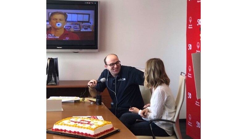 Dan talking with a host while he receives the news of becoming an International Global Ambassador. There is a congratulations cake on the desk.