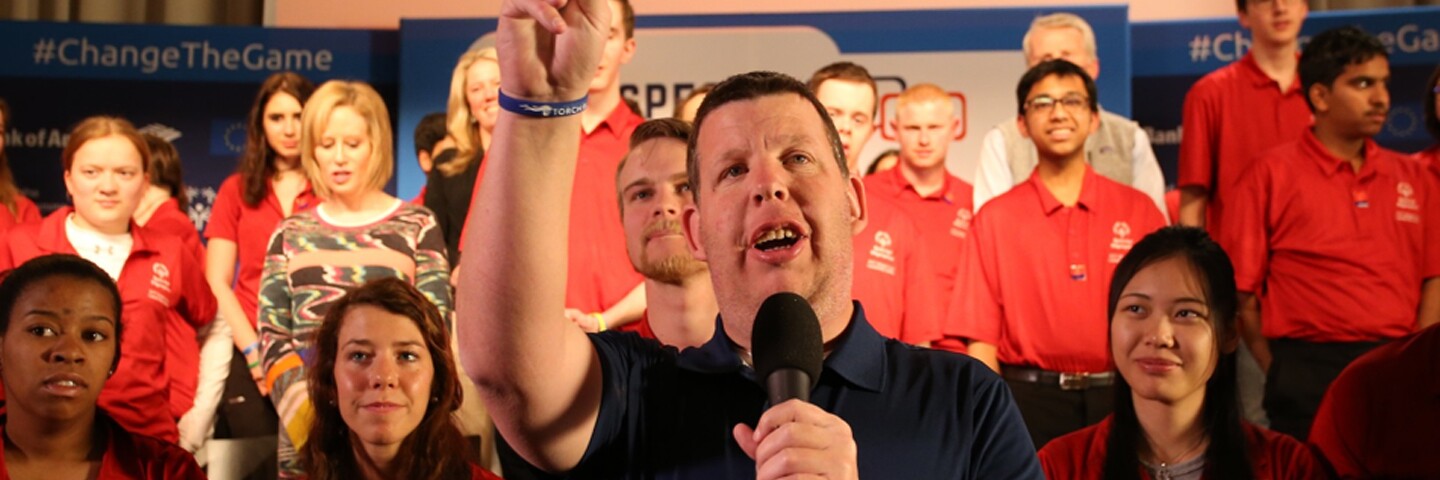 Dustin talking on a mic with athletes stand behind him. 