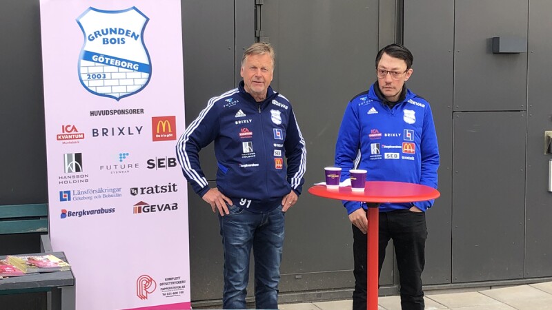 Two men dressed in team gear stand next to a human-sized pink display poster showing the Grunden Bois logo. 