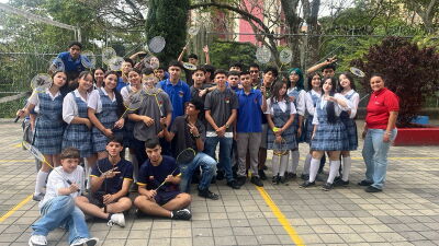 A large group of students gathers outdoors on a school campus holding badminton rackets. Participants pose together on a paved court near a badminton net, with trees and fencing in the background during a youth sports and inclusion activity.