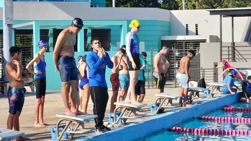 Multiple athletes standing on deck at a swimming pool with two athletes standing on swim block preparing to dive into the pool.