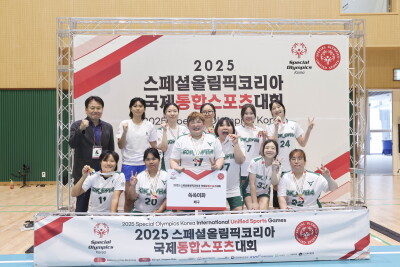 A group of youth leaders pose for a team photo at the 2025 Special Olympics Korea International Unified Sports Games. Participants wearing white and green team jerseys display medals and stand in front of an event backdrop and banner inside an indoor sports venue.