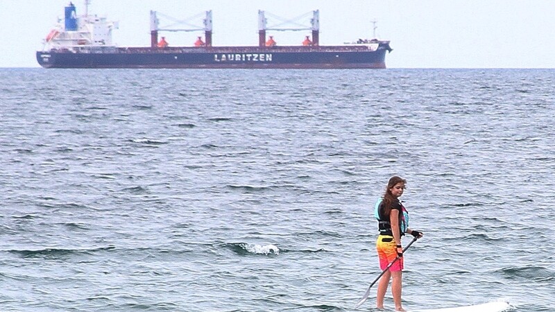 Special Olympics athlete rides paddleboard in ocean with large ship in background. 