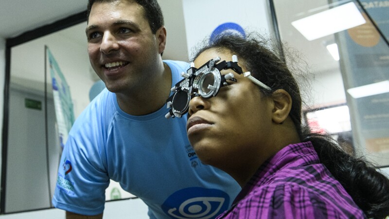 Athlete receiving an eye exam, volunteer standing next to her smiling and looking at results.