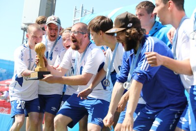A group of men in blue shorts and white t-shirts stand together around one man who is holding a trophy. They have their mouths open as if shouting or cheering.  