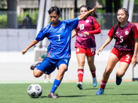 A male player prepares to kick the ball while two women from the opposing team come up behind him. 