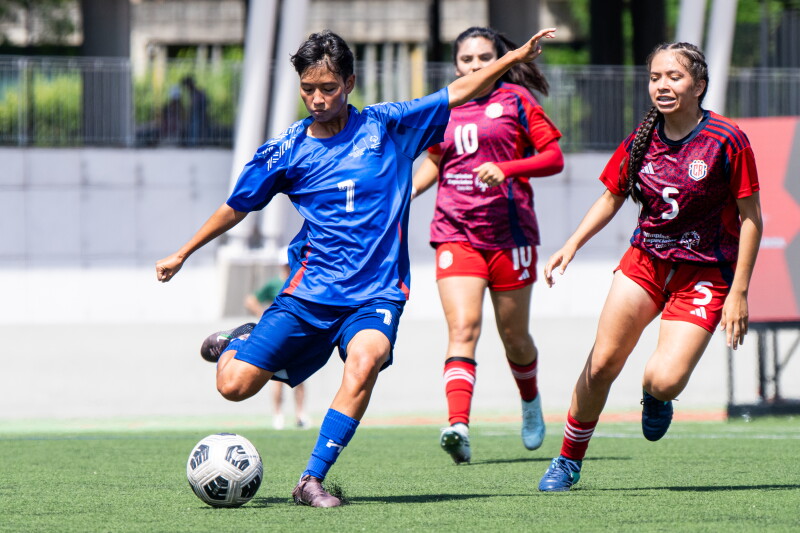 A male player prepares to kick the ball while two women from the opposing team come up behind him. 