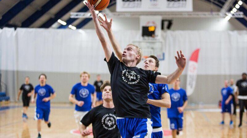 One player in a black play unified t-shirt jumps for a layup shot; a player in a blue play unified t-shirt attempts to block the shot; players behind watch the action and run toward the action. 
