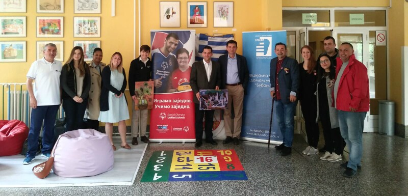 A group of people pose for the camera, standing between Special Olympics branding in a school.