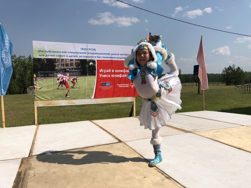 A child on a stage dancing in traditional costume in front of a Special Olympics and Stavros Niarchos Foundation poster. 