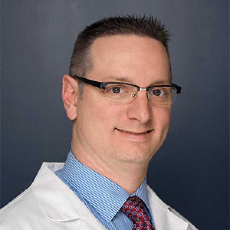 Dr. Peter Seidenberg smiling in a white lab coat with a blue and white pin striped button up and red patterned tie.
