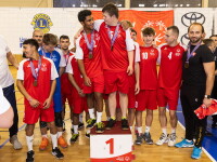 A group of volleyball players in red and white kits stand on and around a first-place podium wearing medals around their necks. 