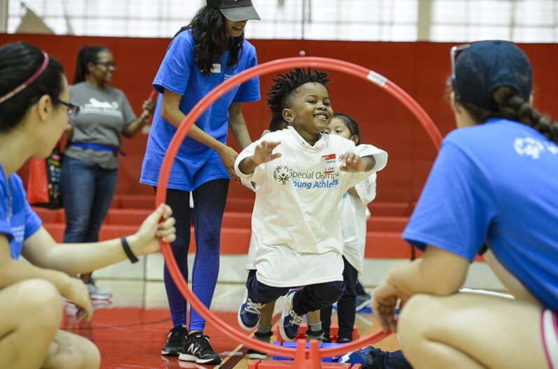 A young athlete smiles as he jumps through a hoop that is being held by two volunteers.