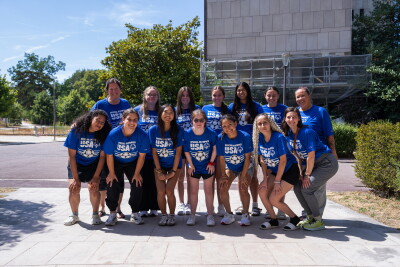 A group of women wearing blue shirts with "USA" in white lettering smile as they pose together outdoors, with a construction facade, trees and shrubs in the background.