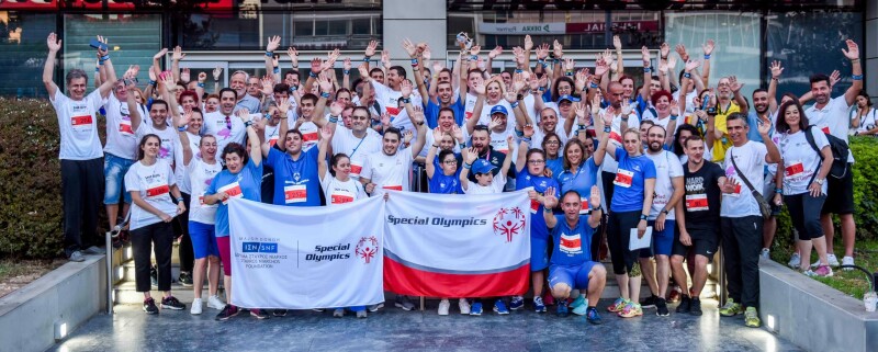 A large group of people in t-shirts and race bibs stand with their hands in the air in celebration behind two banners branded with Special Olympics and SNF logos.