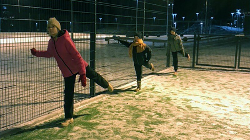 Three young women stand in a row on snow-covered grass, leaning against a fence and stretching their legs out behind them.