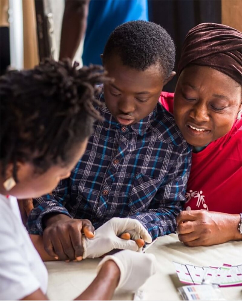 young man sitting on an older woman's lap while a health care provider is pricking his finger for a blood test.