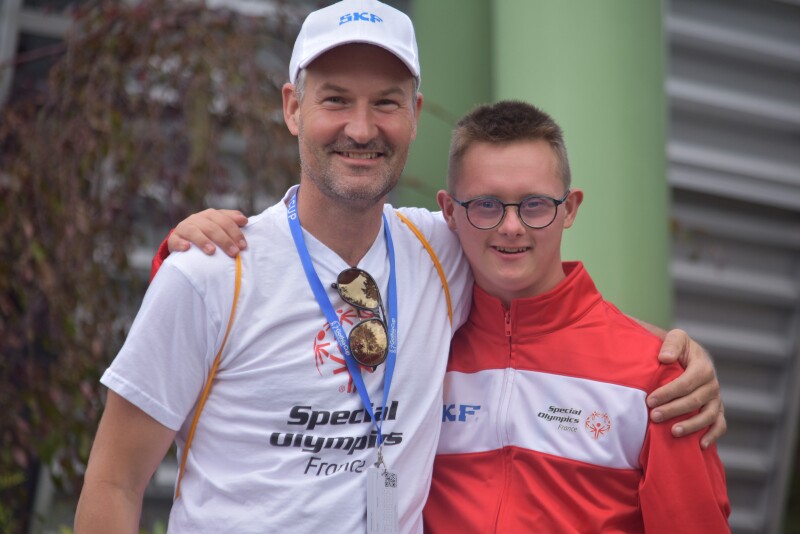 Two men in Special Olympics branded clothing stand facing the camera and smiling with their arms around each other.