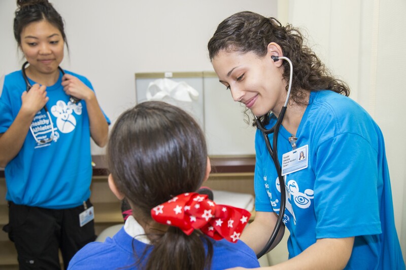 A health professional leans in to listen to a young girl’s heart through a stethoscope.