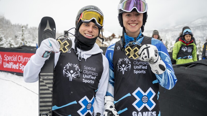 Two skiers standing next to each other proudly display their gold medals while smiling.
