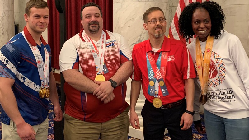 Four people stand and smile in front of the U.S. flag. They are all wearing medals and Special Olympics clothes.