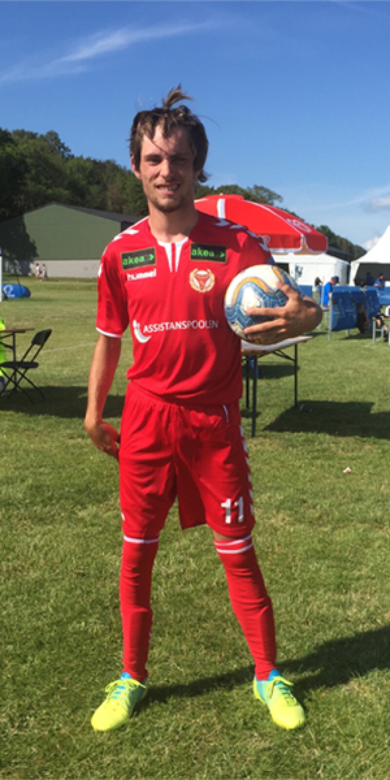 A man stands in a red football kit holding a football and facing the camera at the sidelines of a football pitch.