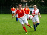 Women in football/soccer jerseys run after a football on a green pitch.