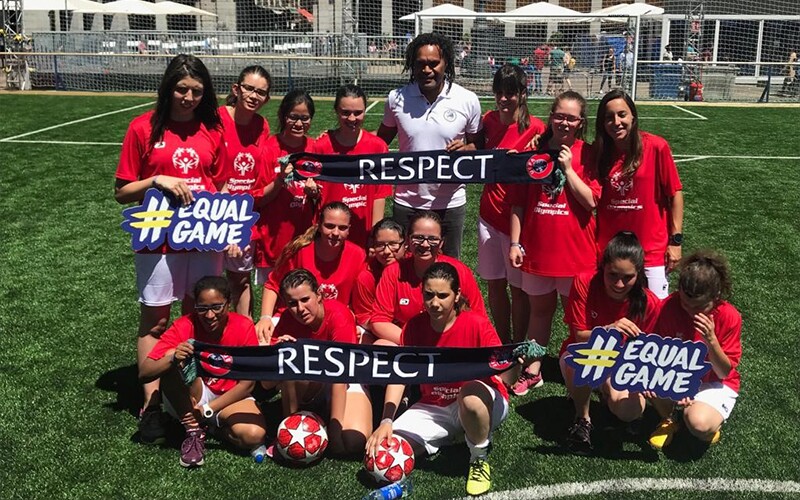 A man stands in the middle of a group of girls in red Special Olympics t-shirts holding signs saying ‘#EqualGame’ and #Respect on a football pitch.