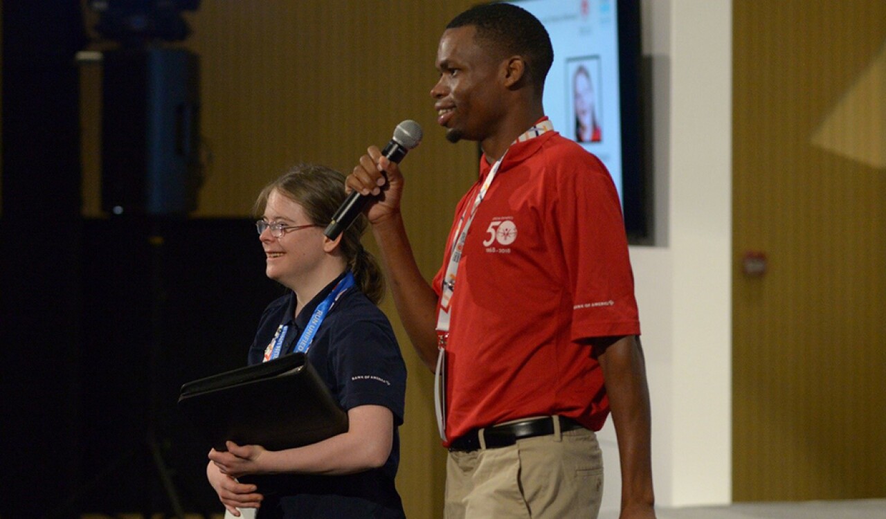 Two young adults standing on stage speaking into a microphone. 