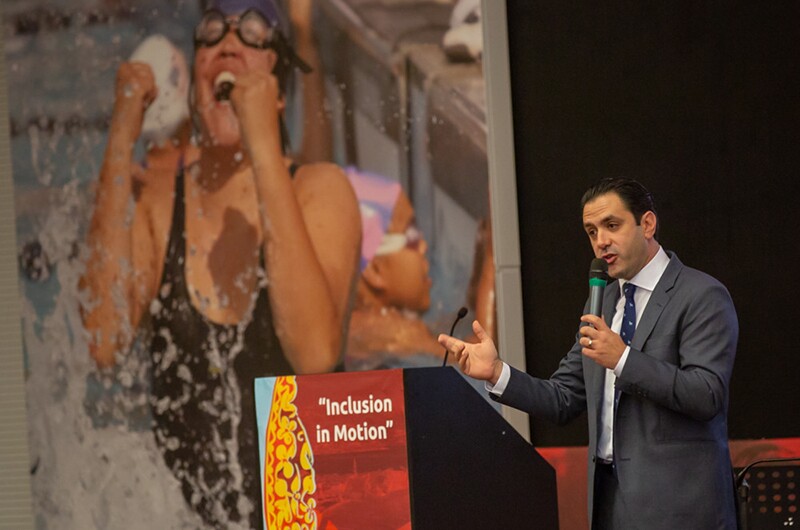 leadership-conferDavid Evangelista, holidng a micraphone and standing behind a podium with text on the front that reads: Inclusion in Motion. He's addresses participants at opening of Special Olympics Europe Eurasia Leadership Conference 2018. A photo in the background is of a young woman splashing out of the water with a look of victory and excitement on her face.