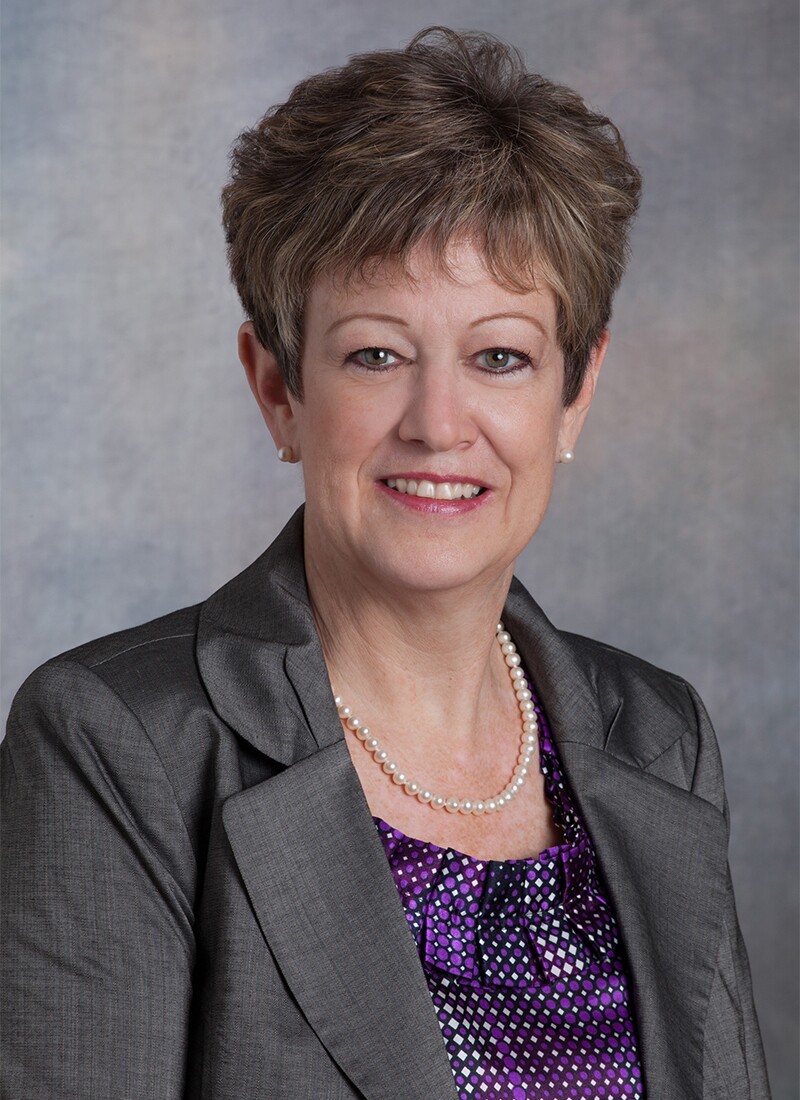 Mary Rosendahl sitting for a professional photo in front of a gray cloudy background. She has short light brown and gray hair, charcoal suit jacket, purple and white diamond-pattern top, and a simple pearl necklace.