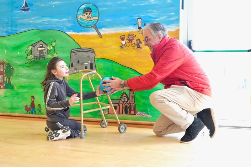 Un hombre con una chaqueta roja se arrodilla en el suelo y estira una pelota hacia un niño que usa un andador. Un niño alcanza una pelota frente a un mural pintado de colores brillantes que representa montañas, edificios, agua y figuras de dibujos animados. 
