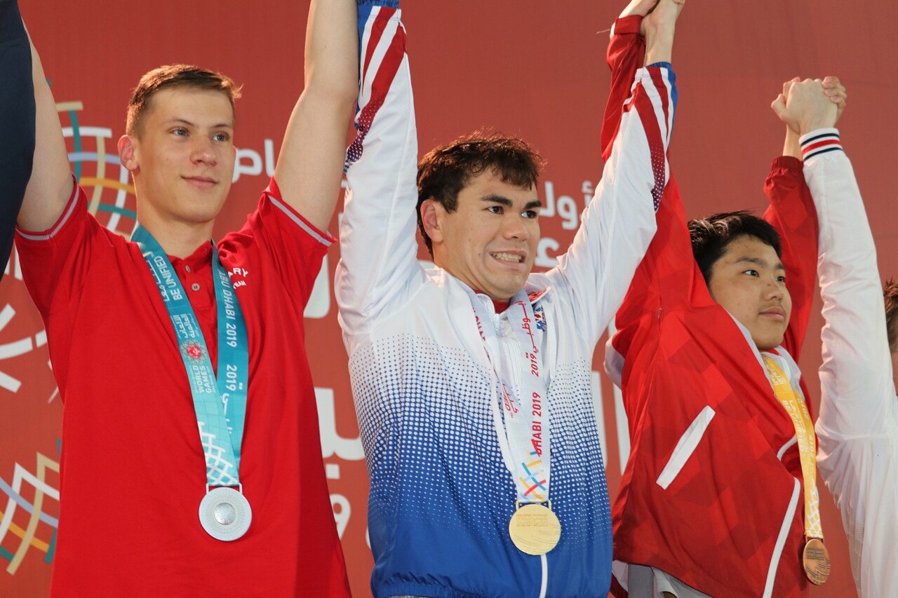 Jonathan placing on the podium with a medal around his neck. An athlete is on either side of him. 