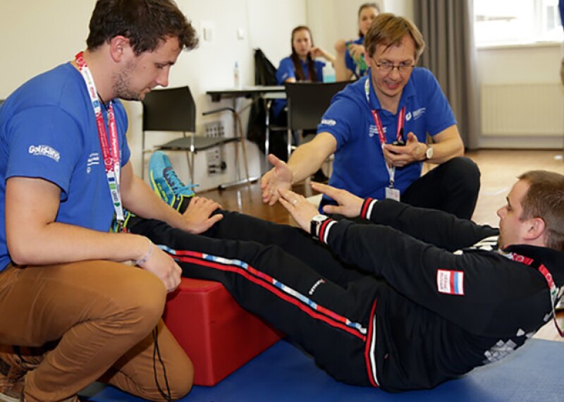 Athlete performing sit-ups during health screening while two volunteers assist and observe.