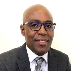 Charles Nyambe portrait. Charcoal gray suite, white shirt, black and white striped tie, black rim glasses.