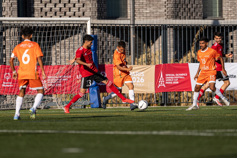 Un jugador con uniforme naranja se estira para patear el balón de un futbolista con camiseta roja cerca del borde del área penal. Dos compañeros de equipo observan de cerca el desarrollo del juego frente a la portería durante un partido fuera de casa.