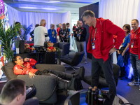 In a busy room full of people, a man in a recliner chair smiles up at a man standing near him.  