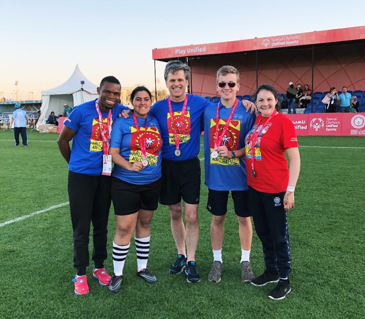 Four young adults and one adult standing in the middle of the soccer pitch in a group with their arms around one another.