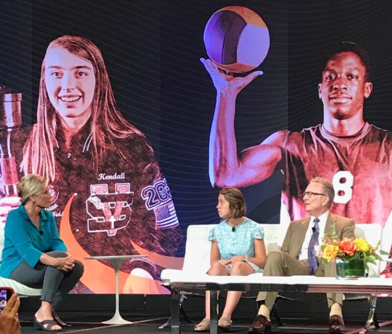 A woman sits facing a young woman and a man on a white sofa with large images of sports players in the background.